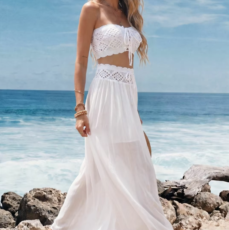 Side view of white crochet-look crop top and flowing sheer maxi skirt set worn by model on coastal rocks.
