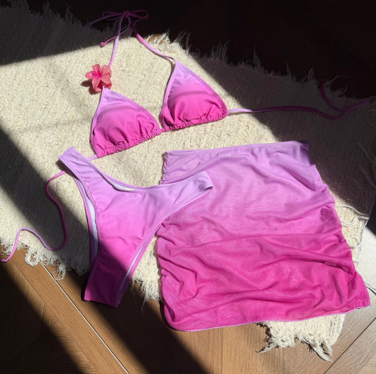 Pink triangle bikini and matching ombré sheer mini skirt set with flower shoulder detail in a flat lay.