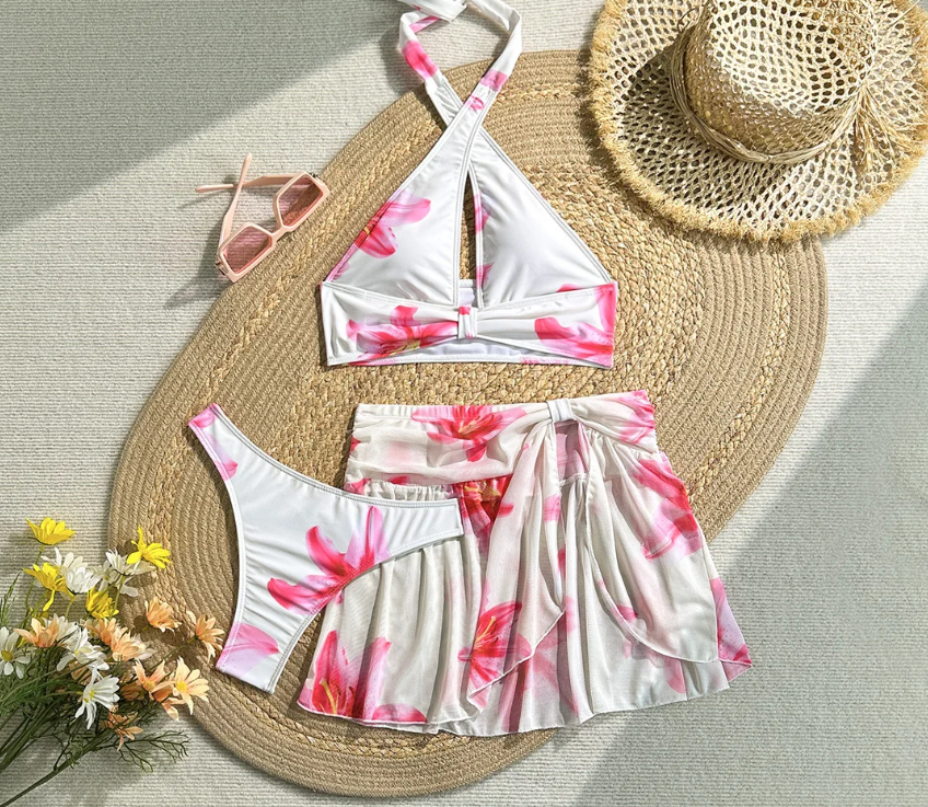 Flat lay of Camellia white floral bikini and matching wrap skirt set with halter top, high-leg bottoms and pink lily print.