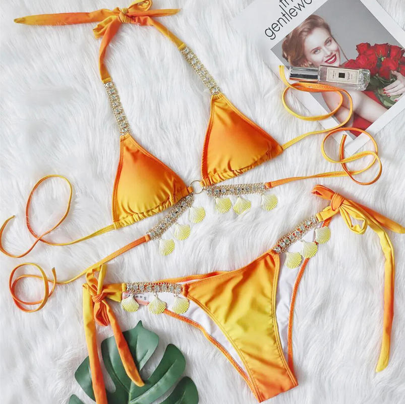 Flat lay of Cancún orange bikini set with triangle halter top, rhinestone trim, shell charms and tie-side bottoms.