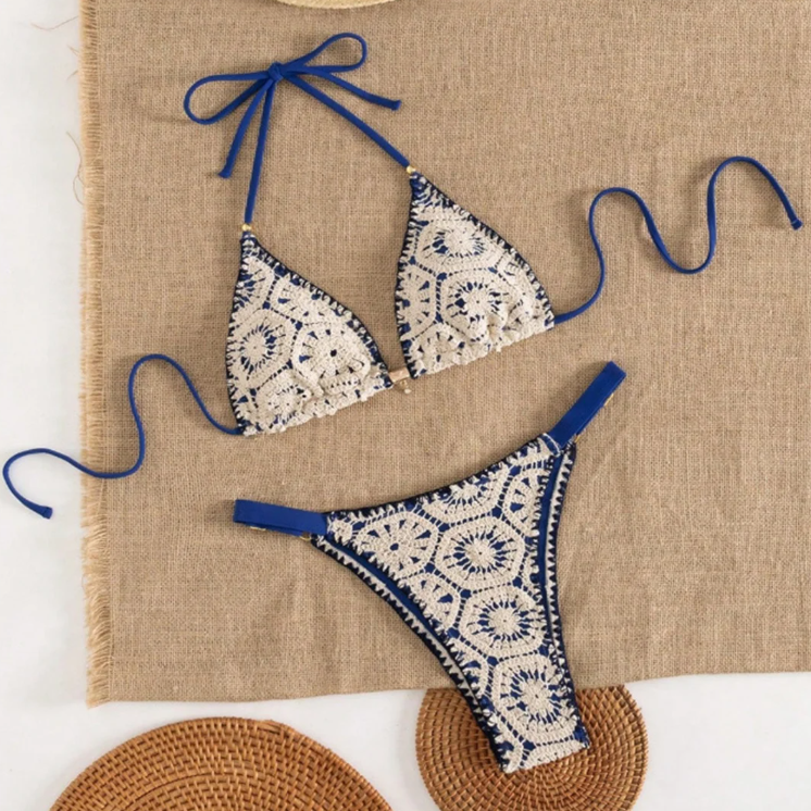Flat lay of blue and ivory crochet-look triangle bikini set with halter ties and high-leg bottoms on a neutral woven backdrop.