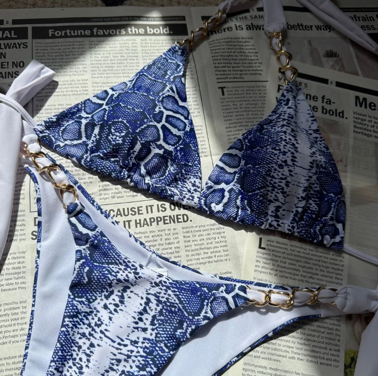 Flat lay close-up of the Delphine bikini set in blue snakeskin print, showing the triangle bikini top and matching bottoms with white ties and gold chain hardware.