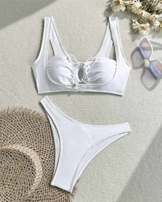 White longline bikini set with gathered lace-up front top and high-leg bottoms arranged in a beach flat lay.