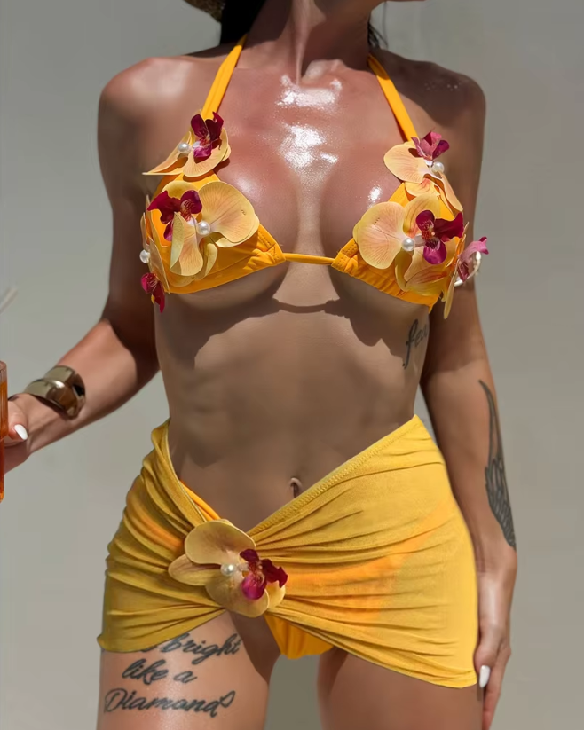 Orange orchid bikini and sheer mini skirt set worn by model with triangle top and floral appliqué details.