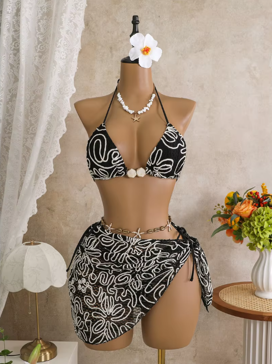 Black corded floral bikini and matching wrap sarong set displayed on mannequin with contrast white embroidery and shell accents.