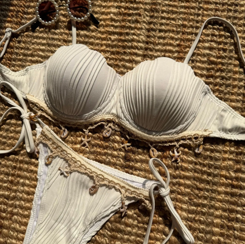 Ivory shell-inspired bikini set flat lay with pleated cups, shell charm trim and tie-side bikini bottoms.