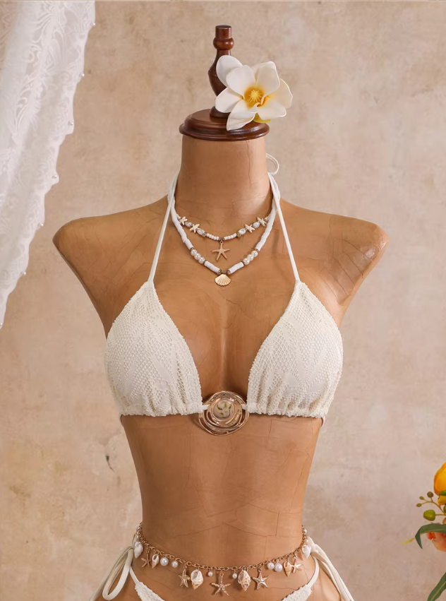 Close-up of ivory openwork triangle bikini top with botanical texture, halter ties and sculptural gold-tone hardware.